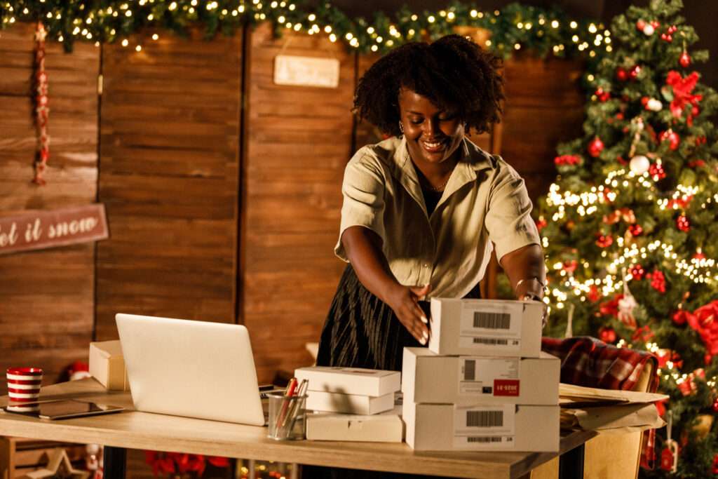 Smiling african woman in white shirt, apron and eyeglasses sitting at decor shop and holding two boxes in hands. Happy saleswoman using modern laptop at work.