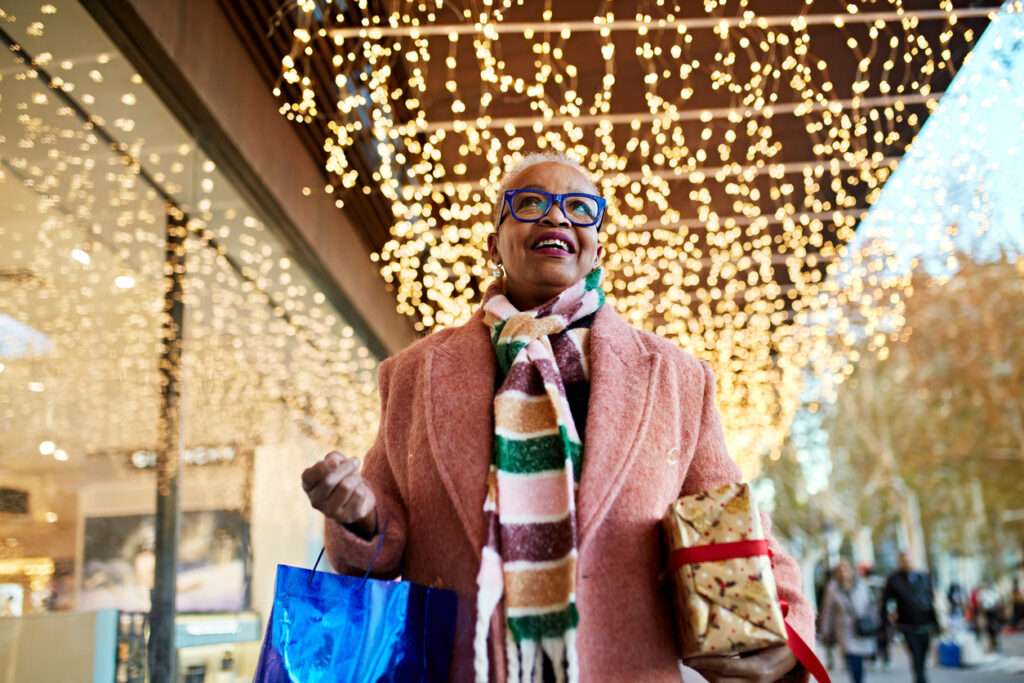 Waist up view of Black woman in late 50s wearing winter coat and scarf, approaching camera in outdoor shopping district beneath glittering lights.